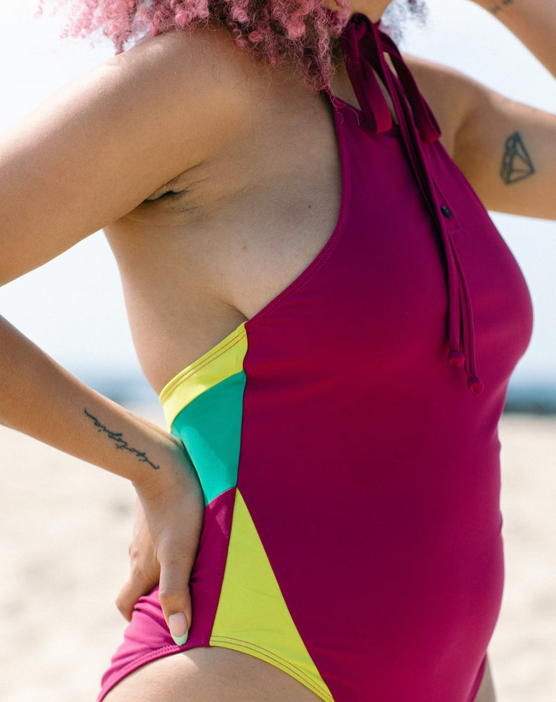 Model showcasing color blocking of lime and emerald green details at the sides and back of the magenta halter one-piece swimsuit she is wearing.