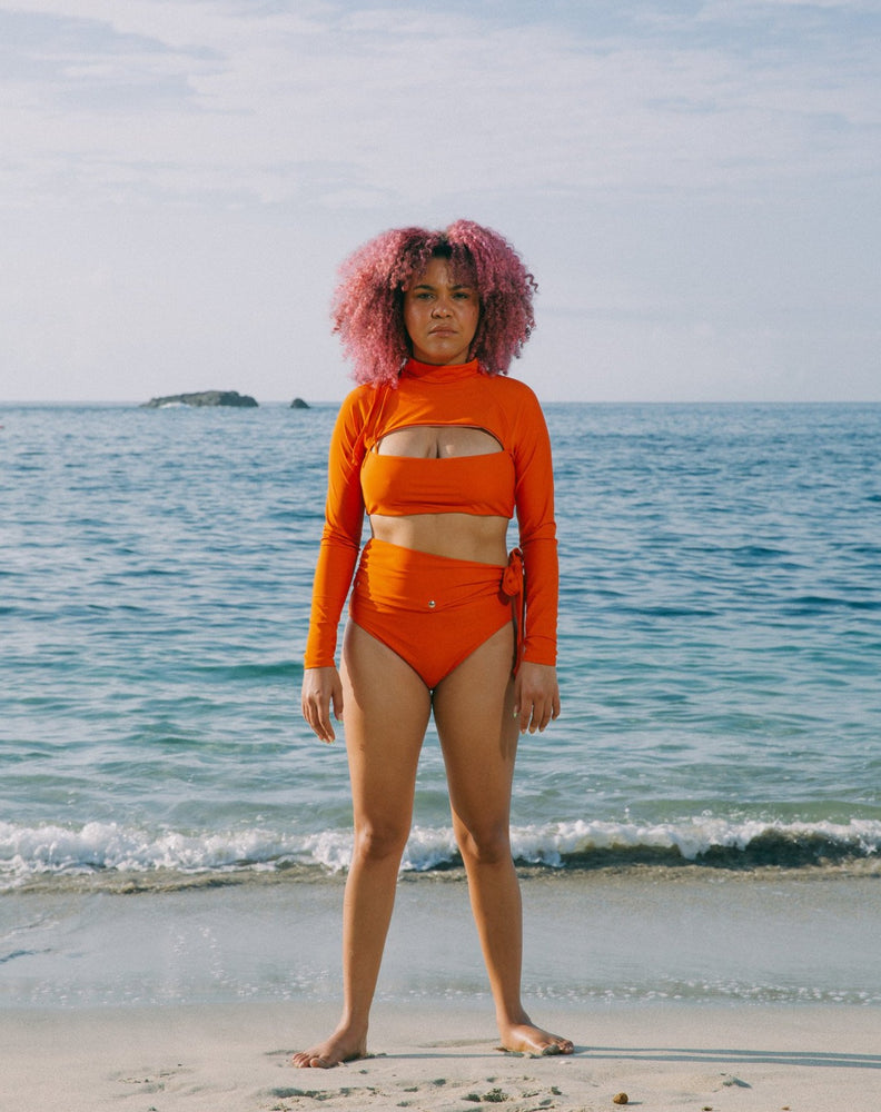Model showcasing how to use zipper pulley of burnt orange long sleeves top with mock neck and cutout at chest. She is also wearing matching burnt orange high-waisted bikini bottom with adjustable snap belt at waist.