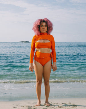 Model with ocean at back, wearing burnt orange high-waisted bikini bottom with snap belt at waist and matching burnt orange long sleeves top with mock neck and cutout at chest.