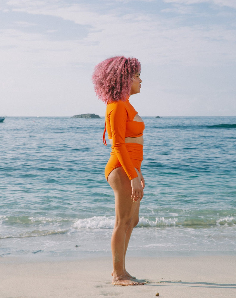 Model with ocean at back looking to the side wearing burnt orange long sleeves top with mock neck and cutout at chest. She is also wearing matching burnt orange high-waisted bikini bottom with adjustable snap belt at waist. 