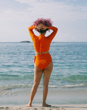 Model looking at sea wearing burnt orange long sleeves top with mock neck and zipper with pulley at back. She is also wearing matching burnt orange high-waisted bikini bottom with adjustable snap belt at waist. 