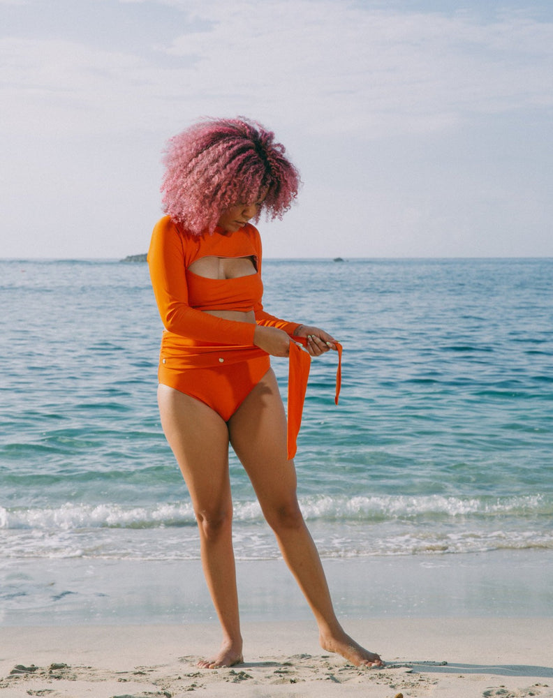 Closeup of model showing her stoma bag in torso, wearing burnt orange high-waisted bikini bottom with unsnapped belt and burnt orange long sleeves top.