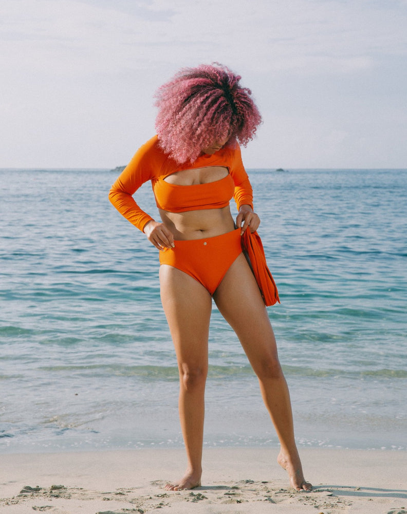 Model with ocean at back adjusting orange high-waisted bikini bottom without snap belt. She is also wearing matching burnt orange long sleeves top with mock neck and cutout at chest.
