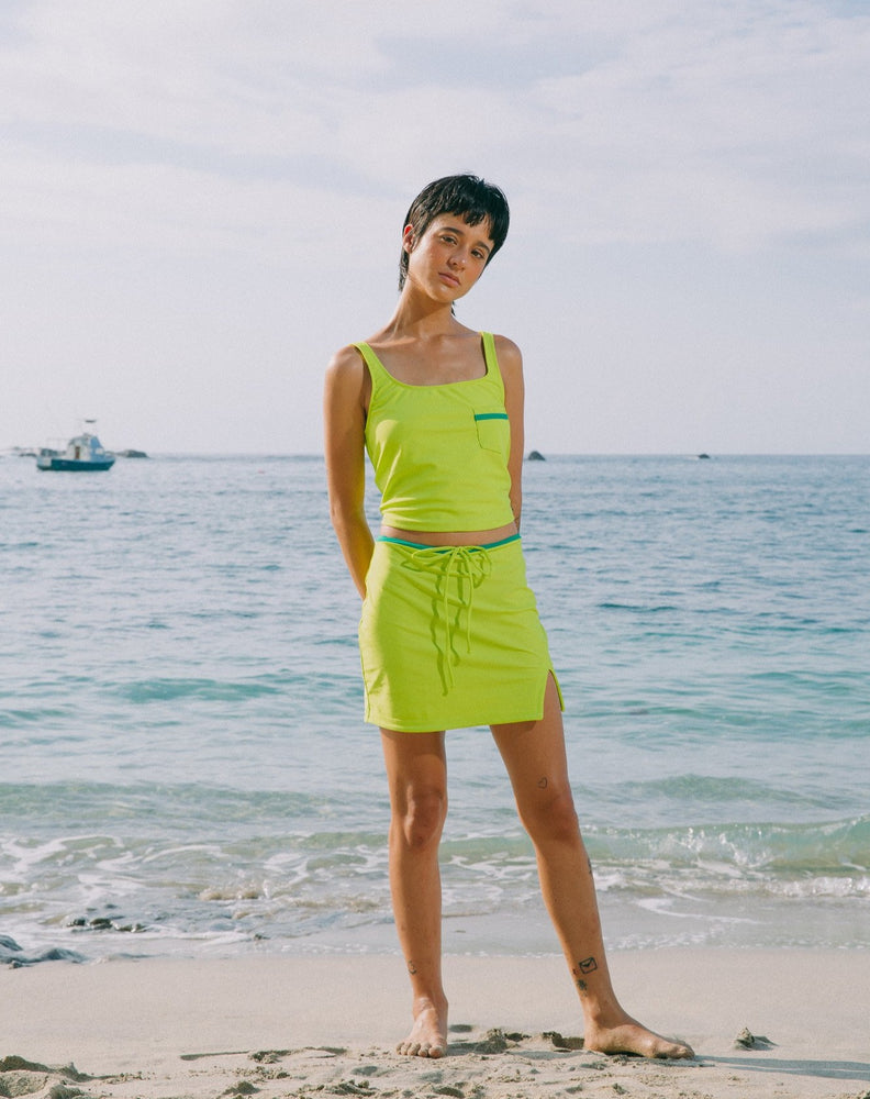 Model showcasing small slit in left side of mid-thigh length lime green skirt with adjustable emerald green string. She is also wearing matching lime green tankini top with scoop neck that comes with a front pocket with emerald green detail.