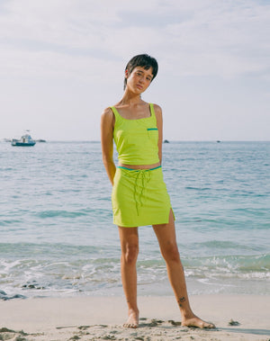  Model with ocean at back wearing mid-thigh length lime green skirt with emerald green waistband and small slit in left side. She is also wearing lime green tankini top with scoop neck that comes with a front pocket with emerald green detail. 