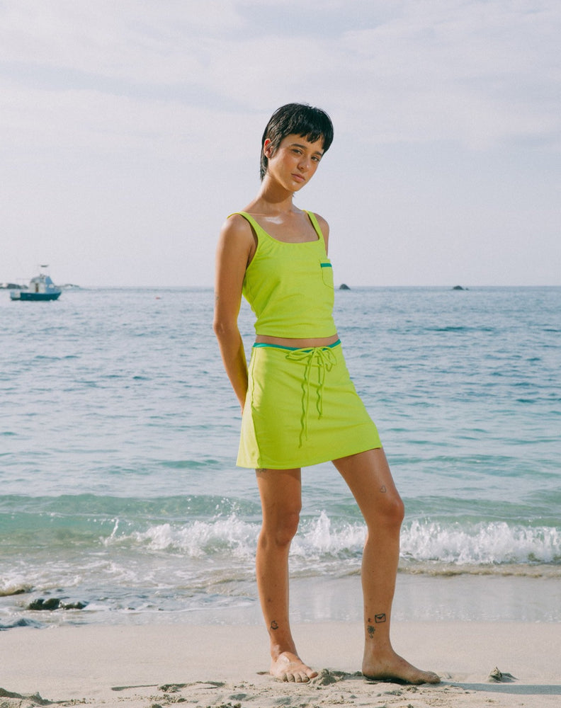 Model with ocean at back wearing mid-thigh length lime green skirt with adjustable emerald green string and small slit in left side. She is also wearing matching lime green tankini top with scoop neck that comes with a front pocket with emerald green detail. 