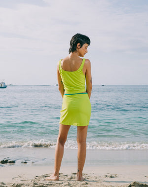 Model with ocean at back, looking to the side, wearing mid-thigh length lime green skirt with adjustable emerald green string and small slit in left side. She is wearing matching lime green tankini top with scoop neck that comes with a front pocket with emerald green detail. 