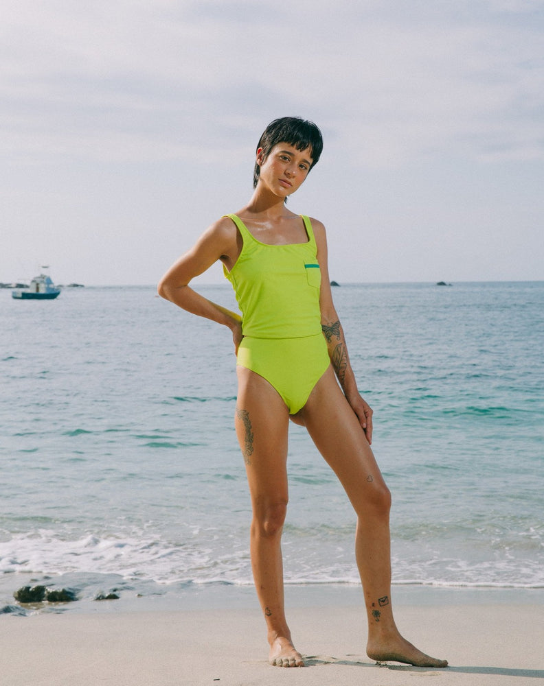 Model with ocean at back wearing lime green tankini top with scoop neck that comes with a front pocket with emerald green detail. She is also wearing matching lime green high-waisted bikini bottom with moderate coverage. 