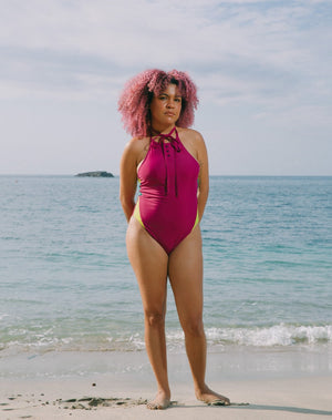 Model with ocean at back wearing magenta halter one-piece swimsuit with lime green details at sides. Decorative button detail at front. Long tying magenta strings for multiple uses.