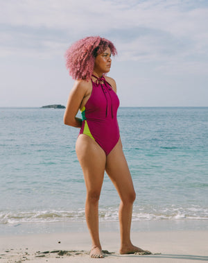 Model looking to side with ocean at back wearing magenta halter one-piece swimsuit with lime green details at sides. Decorative button detail at front. Long tying magenta strings for multiple uses.