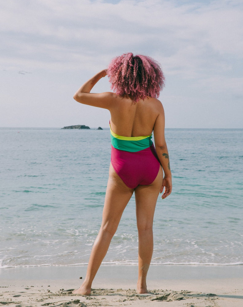 Model looking at ocean with hand in face wearing magenta halter one-piece swimsuit with lime green details at sides. Decorative button detail at front. Long tying magenta strings for multiple uses.