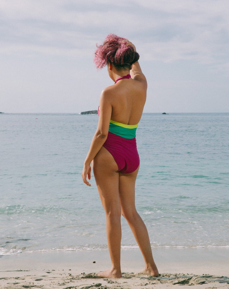 Model looking to side, hair up, with ocean at back wearing magenta halter one-piece swimsuit with lime green details at sides. Decorative button detail at front. Long tying magenta strings for multiple uses.
