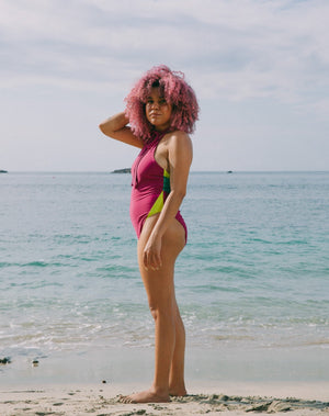 Model to side with hand in hair, ocean at back wearing magenta halter one-piece swimsuit with lime green details at sides. Decorative button detail at front. Long tying magenta strings for multiple uses.
