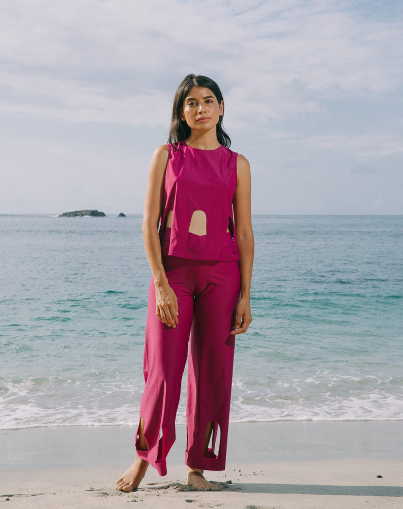 Model with ocean at back wearing magenta high waisted pants with arc cutouts around legs. Slight bell bottom leg. Also wearing magenta color short sleeve top with scoop neck. Arc cutouts around waist. Loose fit.