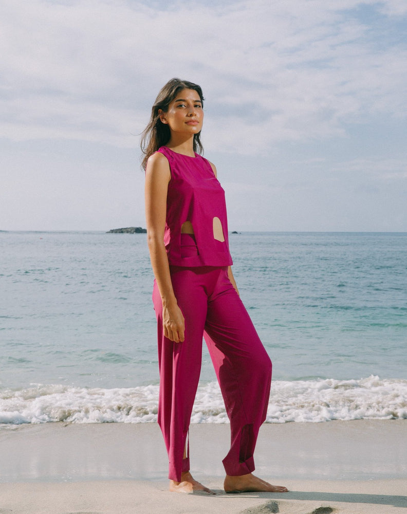 Model with ocean at back wearing magenta color short sleeve top with scoop neck. Arc cutouts around waist. Loose fit. Also wearing matching magenta high waisted pants with arc cutouts around legs. Slight bell bottom leg. 