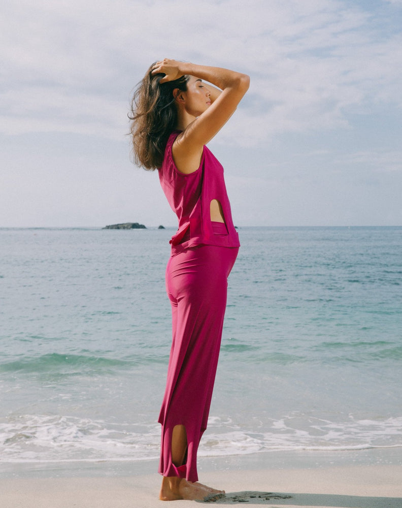 Model with ocean at back, looking to the side with hands on head. She is wearing magenta high waisted pants with arc cutouts around legs. Slight bell bottom leg. Also wearing matching magenta color short sleeve top with scoop neck. Arc cutouts around waist. Loose fit.