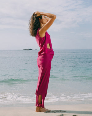Model with ocean at back and hands on head wearing magenta color short sleeve top with scoop neck. Arc cutouts around waist. Loose fit. Also wearing matching magenta high waisted pants with arc cutouts around legs. Slight bell bottom leg. 