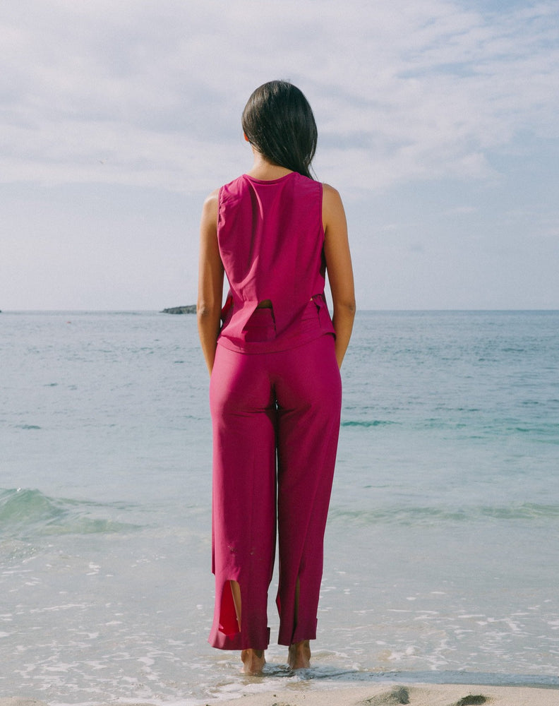 Model facing the ocean is wearing magenta high waisted pants with arc cutouts around legs. Slight bell bottom leg. Also wearing matching magenta color short sleeve top with scoop neck. Arc cutouts around waist. Loose fit.