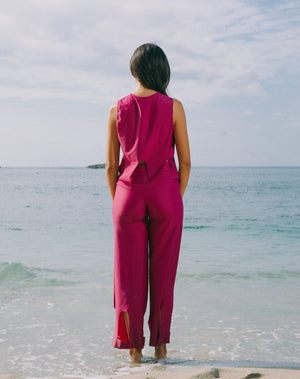 
            
                Load image into Gallery viewer, Model facing the ocean is wearing magenta high waisted pants with arc cutouts around legs. Slight bell bottom leg. Also wearing matching magenta color short sleeve top with scoop neck. Arc cutouts around waist. Loose fit.
            
        