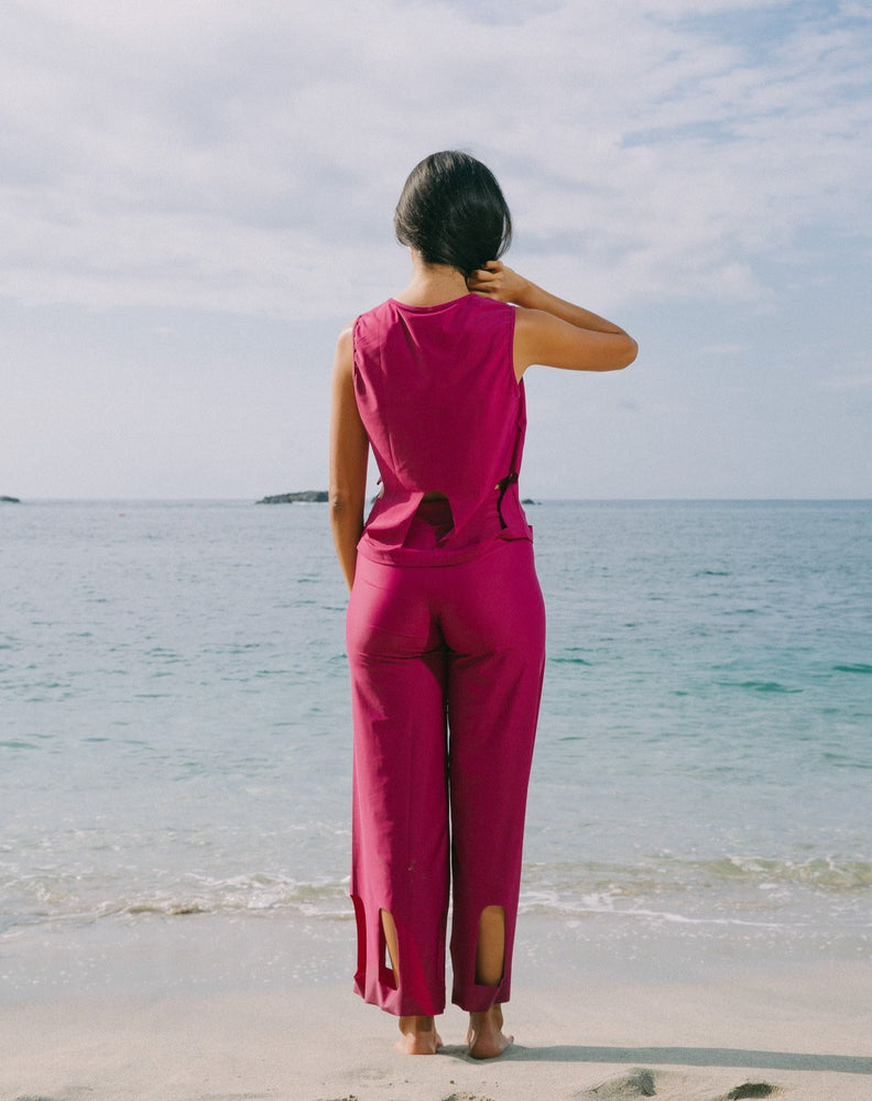 Model looking at ocean and with one hand holding her hair, wearing magenta color short sleeve top with scoop neck. Arc cutouts around waist. Loose fit. Also wearing matching magenta high waisted pants with arc cutouts around legs. Slight bell bottom leg. 